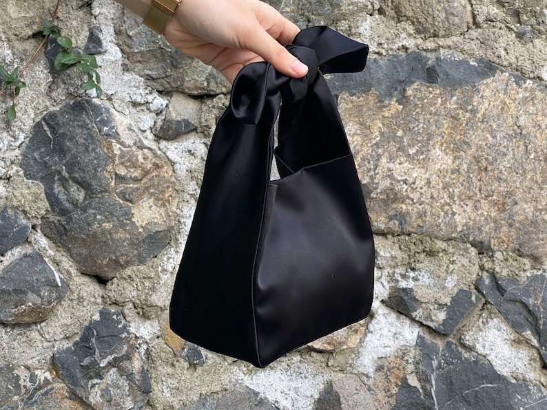 May include: A black satin handbag with a knotted handle. The bag has a rectangular shape with a smooth, shiny texture. The handle is formed by a large knot, creating a stylish and elegant accessory.