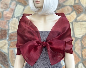 May include: A sheer, deep burgundy organza shawl draped over the shoulders, featuring a large, prominent bow tied at the center front. The fabric is translucent with a subtle, fine-textured weave, creating a structured yet airy appearance.