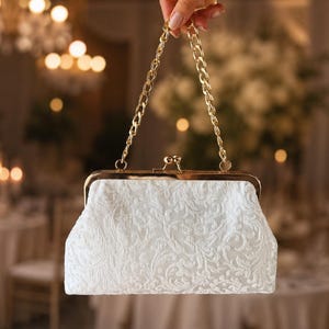 Puede incluir: Un bolso de mano blanco con un intrincado encaje floral superpuesto en tonos a juego, un marco de metal dorado, un cierre de broche y un asa de cadena dorada a juego. El bolso estructurado tiene una forma rectangular con esquinas redondeadas.