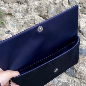 Op de afbeelding: Een zwarte clutch van fluweel met een voering van marineblauw satijn. De clutch heeft een zilveren drukknopsluiting en een afneembare kwast.