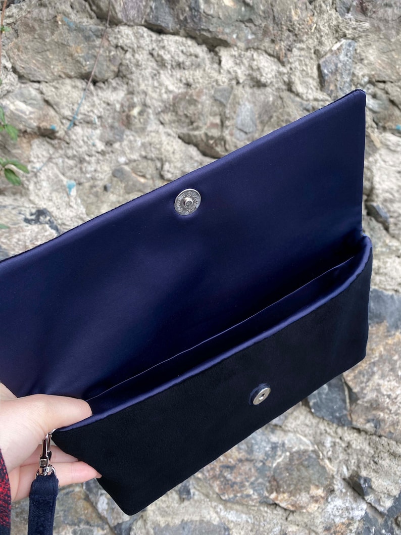 Op de afbeelding: Een zwarte clutch van fluweel met een voering van marineblauw satijn. De clutch heeft een zilveren drukknopsluiting en een afneembare kwast.