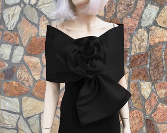 Könnte beinhalten: Ein schwarzes Kleid mit einer großen, dekorativen Schleife und einer rosenförmigen Verzierung. Das Kleid hat ein trägerloses Design und ist aus einem glatten, satinartigen Stoff gefertigt. Der Hintergrund ist eine Steinmauer.