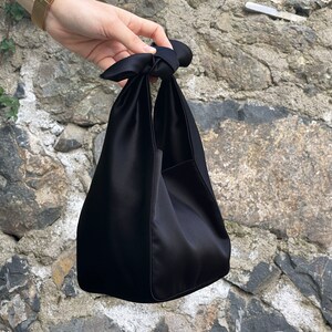 May include: A black satin tote bag with a knotted handle. The bag is folded in half and has a rectangular shape.