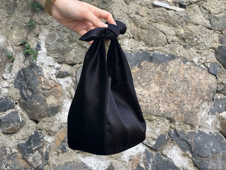 May include: A black satin tote bag with a knotted handle. The bag is folded in half and has a rectangular shape.