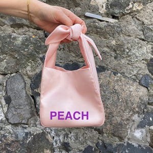 May include: A light pink satin tote bag with a knotted handle. The word "PEACH" is printed in purple on the front of the bag. The bag is held in front of a stone wall.