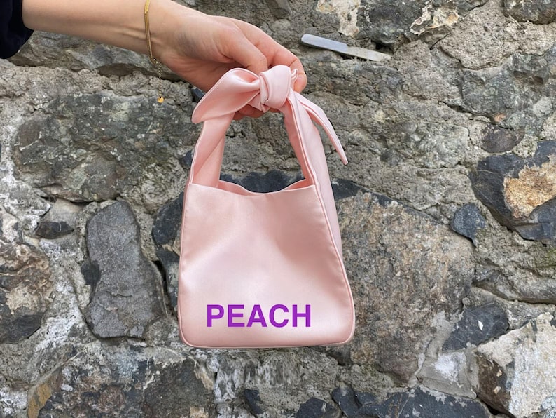 May include: A light pink satin tote bag with a knotted handle. The word "PEACH" is printed in purple on the front of the bag. The bag is held in front of a stone wall.