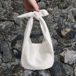 May include: A cream-colored handbag with a knotted handle. The bag has a rounded bottom and a smooth, matte finish. The handle is tied in a decorative knot at the top. The bag is held against a textured stone wall.