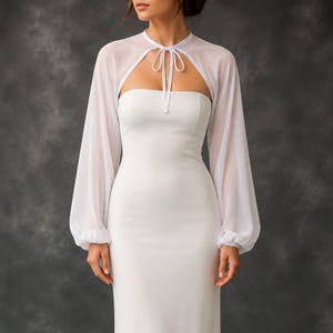 May include: A white strapless gown paired with a matching sheer white bolero. The bolero features long, voluminous bishop sleeves and a delicate tie-front closure at the neckline, creating a layered look over the fitted, floor-length dress.