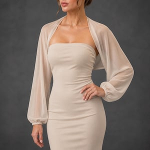 May include: A beige strapless dress paired with a matching sheer bolero jacket featuring long, loose sleeves with gathered cuffs. The garment is made of a solid, smooth fabric and a lightweight, translucent material that drapes over the shoulders.