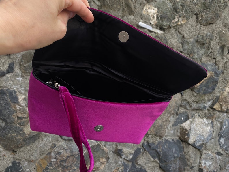Op de afbeelding: Een magenta velvet clutch met een zwart interieur en een magnetische sluiting. De clutch heeft een afneembare polsband.