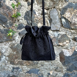May include: A black drawstring bag with a lace-like pattern and a long strap. The bag is hanging against a stone wall.