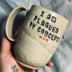 May include: A beige ceramic mug with a large handle. The mug features the text "I AM PLAGUED BY CONCEPTS" in black lettering, with three small red hearts below. The mug has a speckled texture.