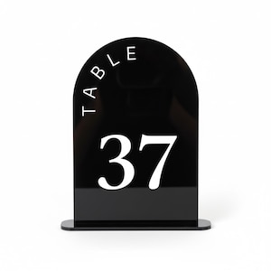 May include: Black acrylic table number with a rounded top. The word "TABLE" is arched across the top in white, with the number "37" prominently displayed below. The base is rectangular.