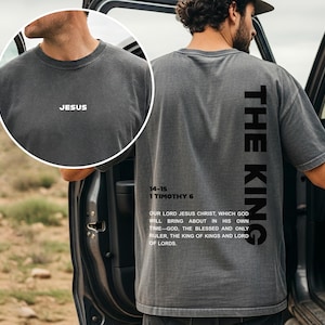 May include: Dark gray t-shirt with the word "JESUS" printed on the front. The back of the shirt features the text "THE KING" vertically, along with a Bible verse. The shirt is a casual, unisex style.