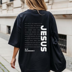 May include: Black oversized t-shirt with the word "JESUS" printed vertically in white on the back. The shirt also features a white block of text with a religious passage from Colossians 1:15-17.