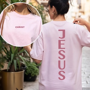 May include: A light pink t-shirt with the word "CHRIST" printed in a darker pink on the front. The back of the shirt features the word "JESUS" printed vertically in a matching darker pink. The shirt is a casual style.