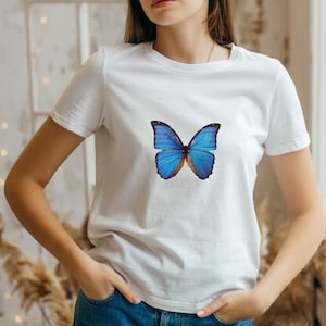 May include: White t-shirt featuring a vibrant blue butterfly graphic. The butterfly has detailed wing patterns with shades of blue and brown. The shirt is a classic crew neck style.