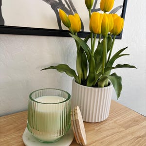 Könnte beinhalten: Dekorative Anordnung mit einer grünen Glaskerze mit Holzdeckel, einem weißen Marmoruntersetzer und einer Topfpflanze mit gelben Tulpen. Die Kerze steht auf einer Holzoberfläche, im Hintergrund ein gerahmter Kunstdruck.