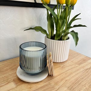 May include: A blue glass candle with a white wax fill sits on a white marble coaster. A wooden lid rests beside the candle. A white ribbed planter with yellow tulips is in the background. The items are on a wooden surface.