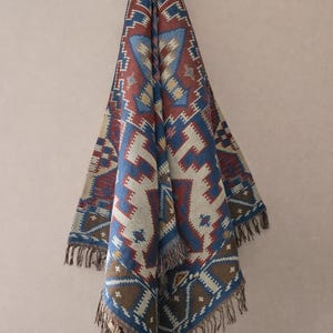 Peut inclure: Une couverture tissée avec un motif géométrique dans les tons de bleu, rouge, marron et crème. La couverture a une bordure à franges et est drapée contre un fond neutre. Le motif présente des formes répétitives.