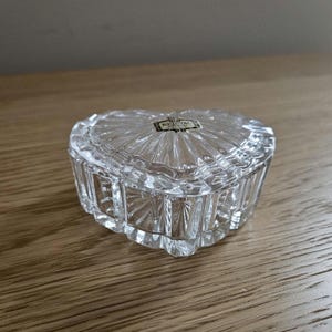 May include: A clear, heart-shaped crystal trinket box with a textured lid and sides. The box has a small rectangular label on the lid. The box is sitting on a wooden surface.
