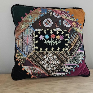 May include: Square decorative pillow with a patchwork design. The center features a black panel with embroidered flowers. The patchwork includes various fabrics in shades of brown, burgundy, and blue, with embroidered details. The pillow has a black velvet border.