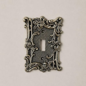 Könnte beinhalten: Eine rechteckige Lichtschalterabdeckung mit antikem Messing-Finish. Die Oberfläche weist eine texturierte, punktierte Mitte und einen erhabenen, verzierten Rand auf, der mit skulpturalen Rosenmotiven und geschwungenen Ranken verziert ist.