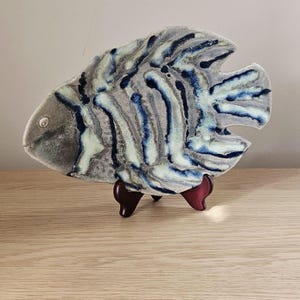 May include: A ceramic fish-shaped decorative plate with a gray, blue, and white striped pattern. The plate is displayed on a dark wooden stand, showcasing its unique design and artistic appeal. The fish plate is a decorative item.