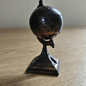 Pode incluir: Um pequeno apontador de lápis em forma de globo de metal escuro, com um design de mapa-múndi esférico montado numa base quadrada decorativa com detalhes ornamentados. O objeto tem um acabamento envelhecido e inclui um orifício lateral.