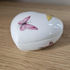 May include: A heart-shaped, white ceramic trinket box with a lid. The lid is decorated with pink and yellow butterfly illustrations. The box is sitting on a wooden surface.