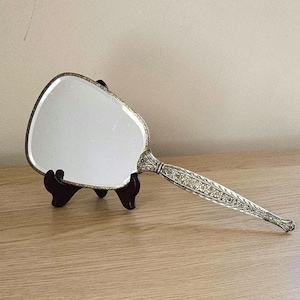 May include: A vintage hand mirror featuring a silver-toned handle and frame. The mirror has an oval shape and is supported by a small, dark wooden stand. The handle is decorated with floral patterns.