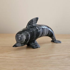 May include: A marble dolphin figurine in shades of gray and white. The sculpture features a sleek, curved body, a dorsal fin, and a pointed snout. The dolphin is posed as if swimming, with its tail and flippers extended. The figurine is displayed on a wooden surface.
