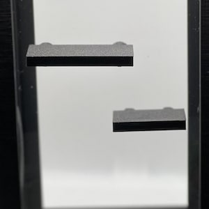 May include: A black display case with two small, dark grey shelves. The shelves are at different heights inside the case. The case has a minimalist design and is set against a white background.