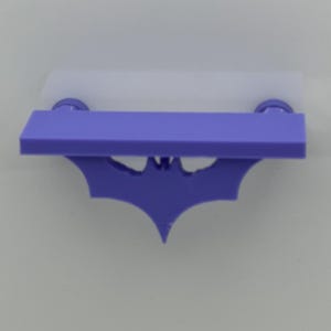 May include: A purple shelf with a bat symbol cutout. The shelf has a rectangular shape with rounded edges and two circular supports. A white, rectangular piece is attached to the top of the shelf.