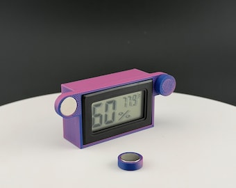 May include: A purple and blue rectangular digital hygrometer with a black screen displaying 60% humidity and 77.9 degrees Fahrenheit. The device features two circular side mounts with silver magnets and includes a separate small circular magnet.