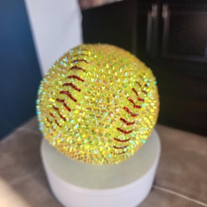 May include: A yellow softball covered in iridescent rhinestones with red stitching. The softball is displayed on a white, round pedestal. The softball is a decorative item.