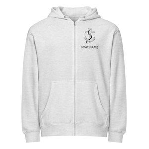 Puede incluir: Una sudadera con capucha gris claro con cremallera, un gráfico de ancla negra y el texto "BOAT NAME". La sudadera con capucha tiene una capucha, una cremallera de largo completo y dos bolsillos delanteros. El gráfico del ancla está en el pecho superior izquierdo.