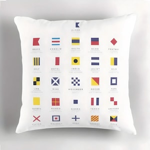 May include: White square cushion featuring a collection of nautical signal flags. Each flag represents a letter of the alphabet, with the letter name printed below. The flags are in various shapes and colours, including red, blue, yellow, and black.