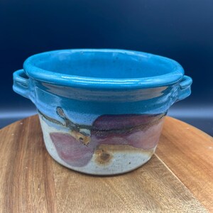 May include: A small ceramic bowl with two side handles, featuring a vibrant turquoise blue glaze on the upper portion and a speckled off-white base decorated with abstract, muted purple and brown painted shapes around the exterior.