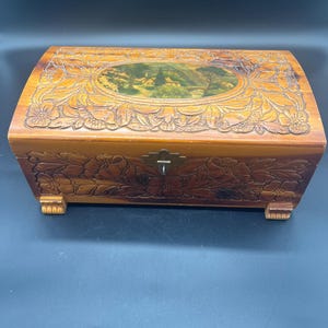 May include: A wooden jewelry box with a landscape painting on the lid. The box is rectangular with rounded edges and is adorned with carved floral patterns. It has a small metal clasp and rests on four small feet.