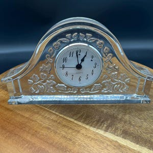 May include: A clear glass quartz clock with a silver clock face. The clock has a decorative floral design and is set on a wooden surface. The clock face displays the time as a few minutes after 12 o'clock.