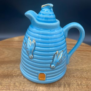 May include: A light blue ceramic creamer shaped like a beehive, with horizontal ridges and a handle. The creamer has gold accents on the spout, lid, and decorative bee-like elements.