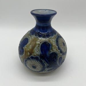May include: A round ceramic vase with a narrow neck and flared rim. The body features a mottled tan and brown glaze with embossed dark blue floral patterns and swirling leaf designs. The neck and rim are finished in a solid, glossy dark blue glaze.