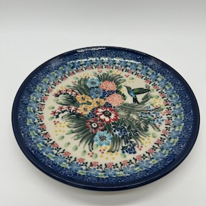 May include: A round ceramic plate featuring a dark blue rim with a decorative pattern. The center displays a colorful floral arrangement with red, yellow, and blue flowers, green foliage, and a small hummingbird in flight against a cream background.