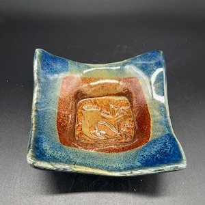May include: A square ceramic dish with a raised center and curved edges. The dish features a blue rim, a reddish-brown middle section, and a textured, brown center with a carved design. The dish is likely handmade.