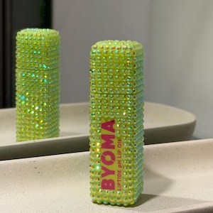 May include: A green lip oil bottle encrusted with iridescent rhinestones. The bottle features the pink text "BYOMA LIPTIDE PH LIP OIL". A mirror reflects the bottle, showing a second lip oil bottle.