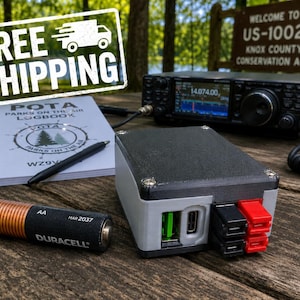 May include: A black and grey rectangular power distribution box with a USB-C port, a green USB-A port, and red and black power terminals. Next to it is a Duracell AA battery, a spiral-bound POTA logbook with a pen, and a black amateur radio transceiver.