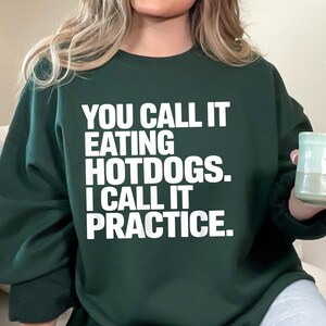 Op de afbeelding: Donkergroene sweatshirt met de tekst "YOU CALL IT EATING HOTDOGS. I CALL IT PRACTICE." in witte blokletters. De sweatshirt heeft een ronde hals en lange mouwen. De persoon op de afbeelding draagt de sweatshirt.
