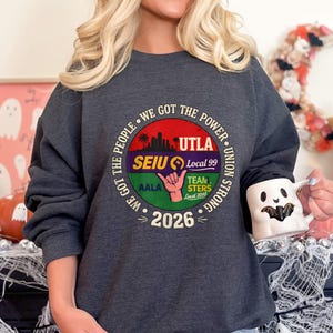 May include: Dark gray sweatshirt with a circular graphic. The graphic includes the text "WE GOT THE POWER" and "UTLA SEIU Local 99 UNION STRONG" with a hand symbol and the year "2026". The sweatshirt is a crew neck style.