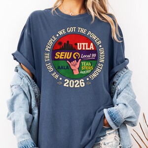 May include: A navy blue t-shirt with a circular graphic featuring the text "WE GOT THE POWER" and union logos. The graphic includes the text "UTLA", "SEIU Local 99", "TEAMSTERS Local 2010", and "2026". The shirt has rolled-up sleeves.